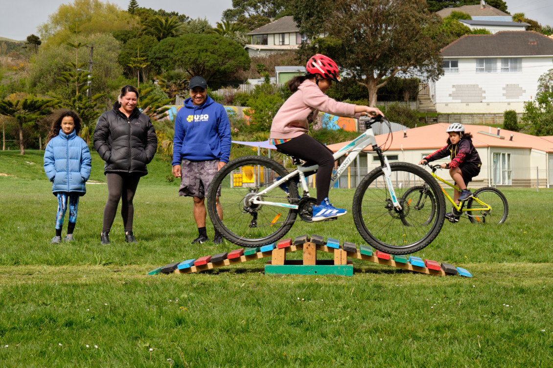 A girl bikes over a small ramp while her family smiles and watches